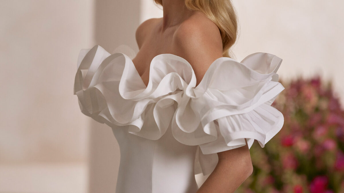 Vestido de noiva de corte direito e estilo juvenil, confecionado em crepe e tafetá. O modelo é completado com um decote envolvente de folhos. Um exclusivo modelo Rosa Clará Couture.