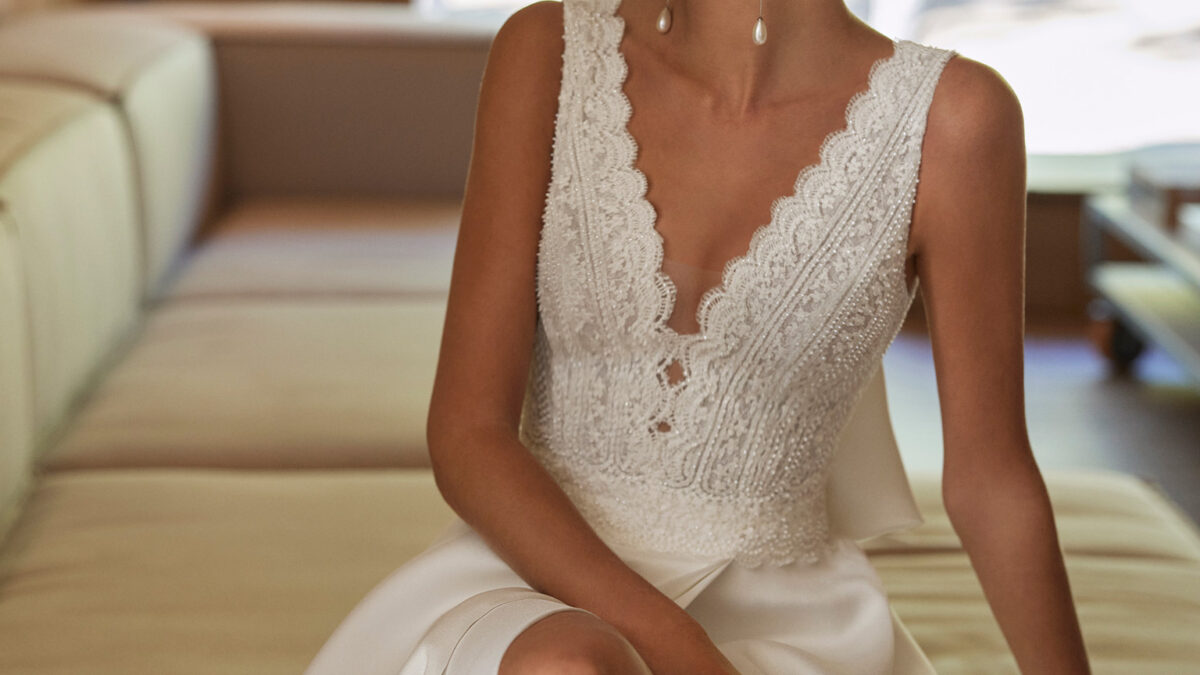 Vestido de noiva de corte em A e estilo clássico, confecionado em Sienna, renda e brilhantes. Descubra um decote profundo, umas costas em V, uma saia com abertura lateral e um laço de adorno nas costas. Um fato de noiva Rosa Clará.