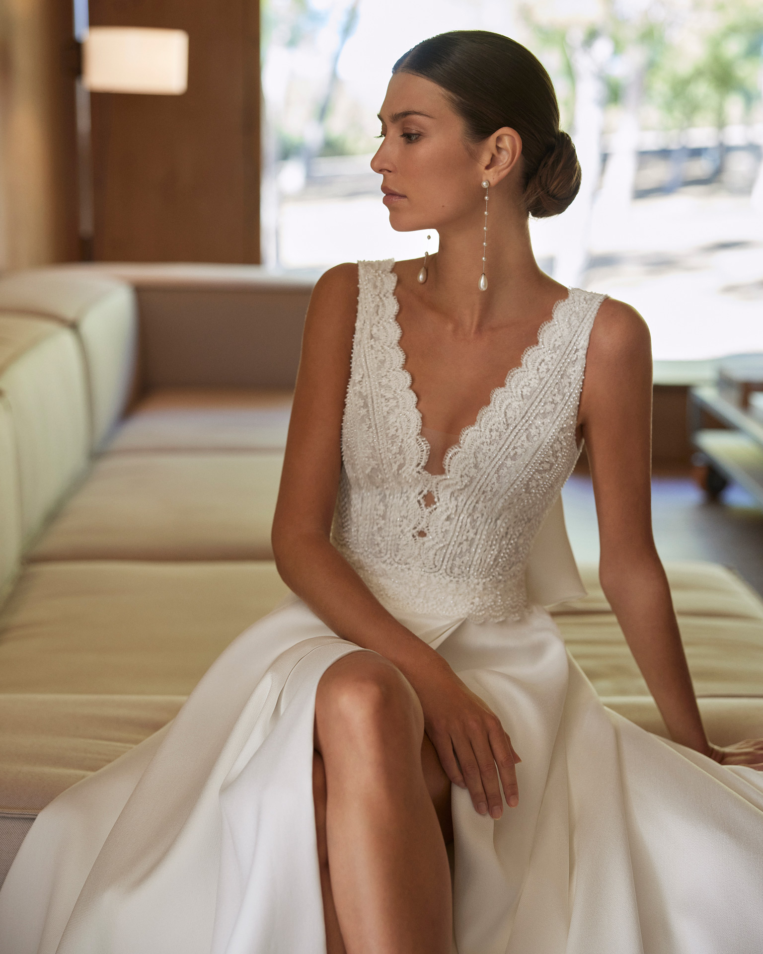 Vestido de noiva de corte em A e estilo clássico, confecionado em Sienna, renda e brilhantes. Descubra um decote profundo, umas costas em V, uma saia com abertura lateral e um laço de adorno nas costas. Um fato de noiva Rosa Clará.