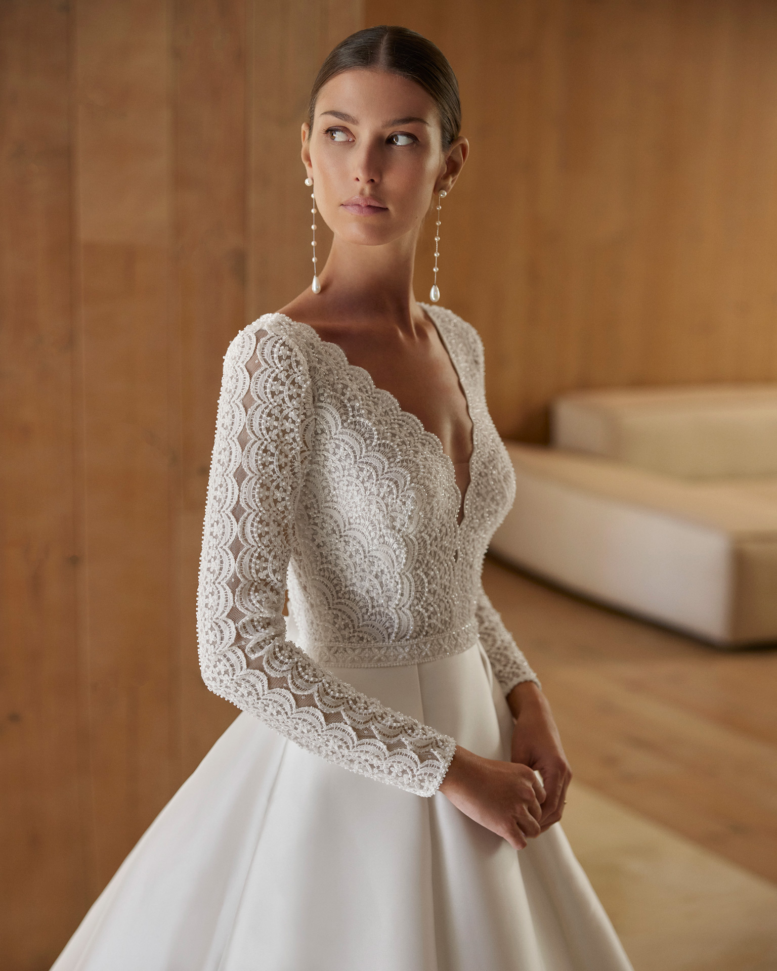 Vestido de noiva de corte em A e estilo clássico, confecionado em mikado combinado com renda e brilhantes. Descubra um decote profundo, umas costas com abertura e manga comprida. Completa-se com uma flor de adorno. Um delicado modelo Rosa Clará.