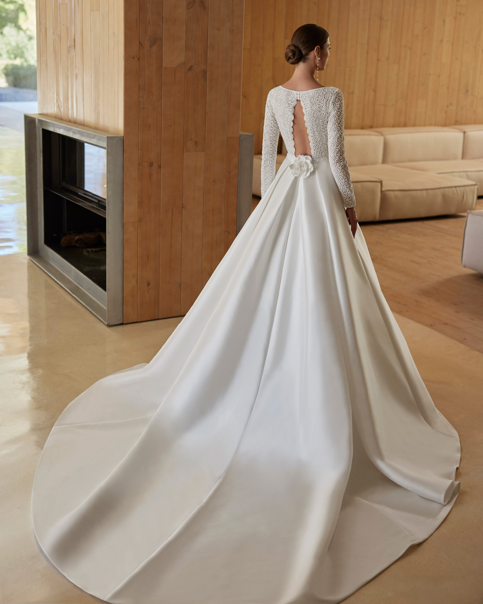 Vestido de noiva de corte em A e estilo clássico, confecionado em mikado combinado com renda e brilhantes. Descubra um decote profundo, umas costas com abertura e manga comprida. Completa-se com uma flor de adorno. Um delicado modelo Rosa Clará.