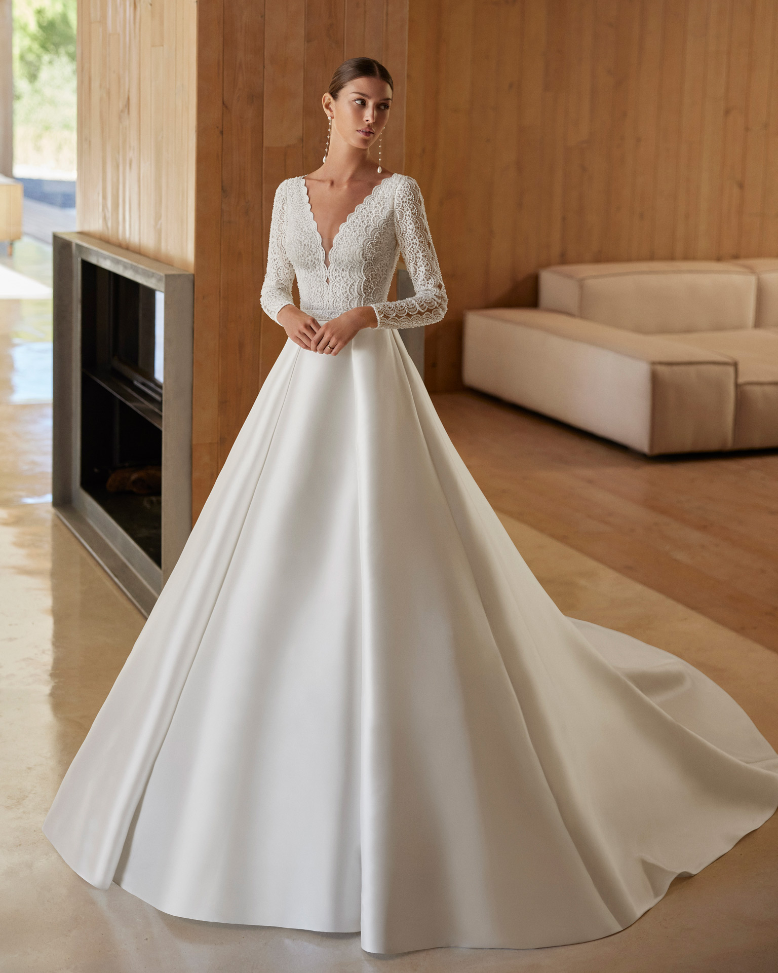 Vestido de noiva de corte em A e estilo clássico, confecionado em mikado combinado com renda e brilhantes. Descubra um decote profundo, umas costas com abertura e manga comprida. Completa-se com uma flor de adorno. Um delicado modelo Rosa Clará.