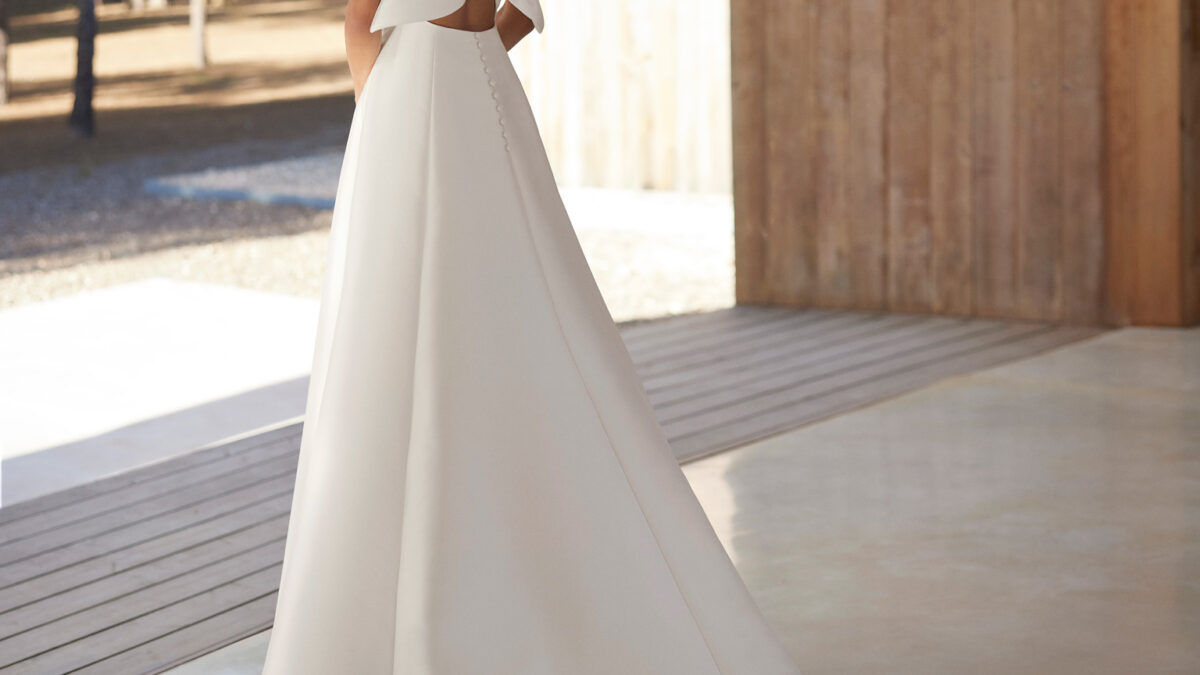 Vestido de noiva confecionado em mikado, com decote profundo e laço de adorno nas costas. Completa-se com alças, uma saia com abertura lateral e uma flor de adorno. Um modelo de sonho Rosa Clará.