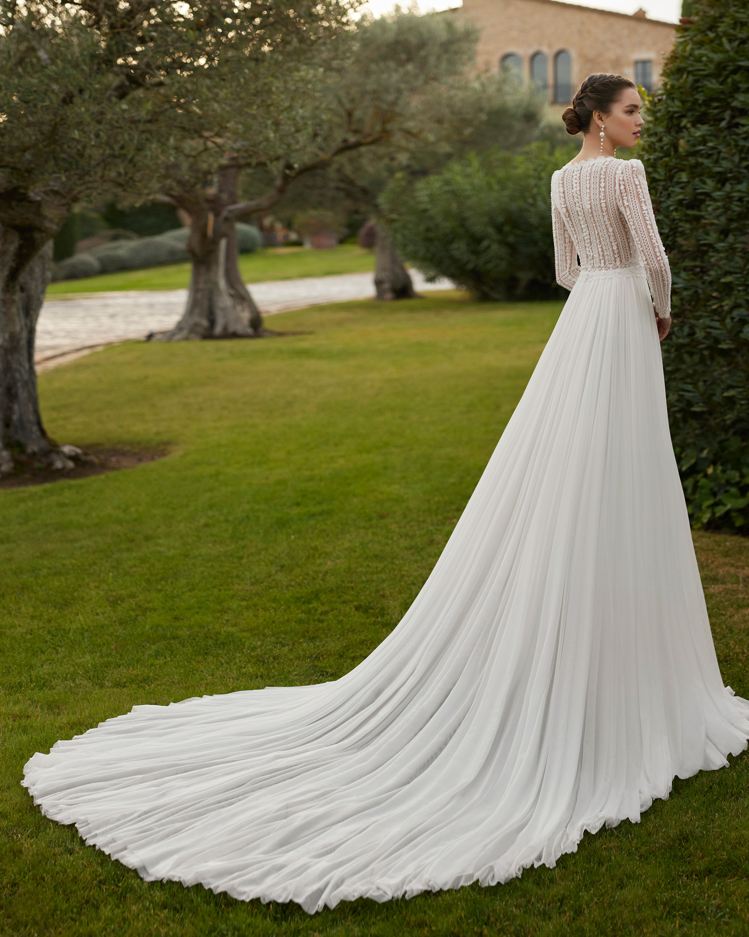 Vestido de noiva comprido, de corte reto e estilo boémio, confecionado com renda e brilhantes e saia em muss. Completa-se com um decote em V, costas fechadas e manga comprida.