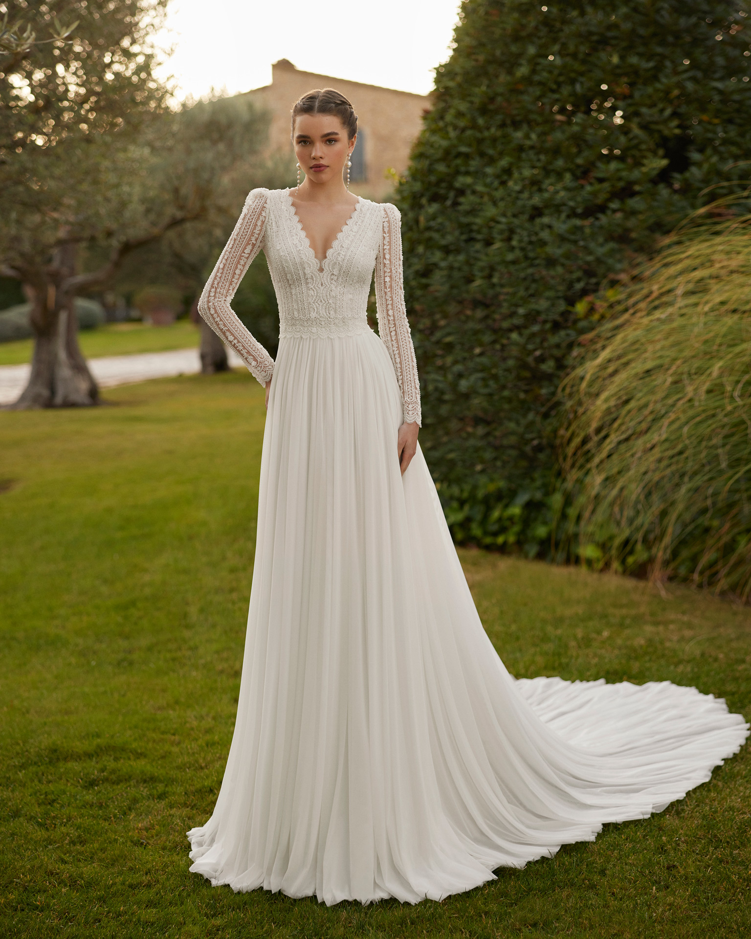 Vestido de noiva comprido, de corte reto e estilo boémio, confecionado com renda e brilhantes e saia em muss. Completa-se com um decote em V, costas fechadas e manga comprida.