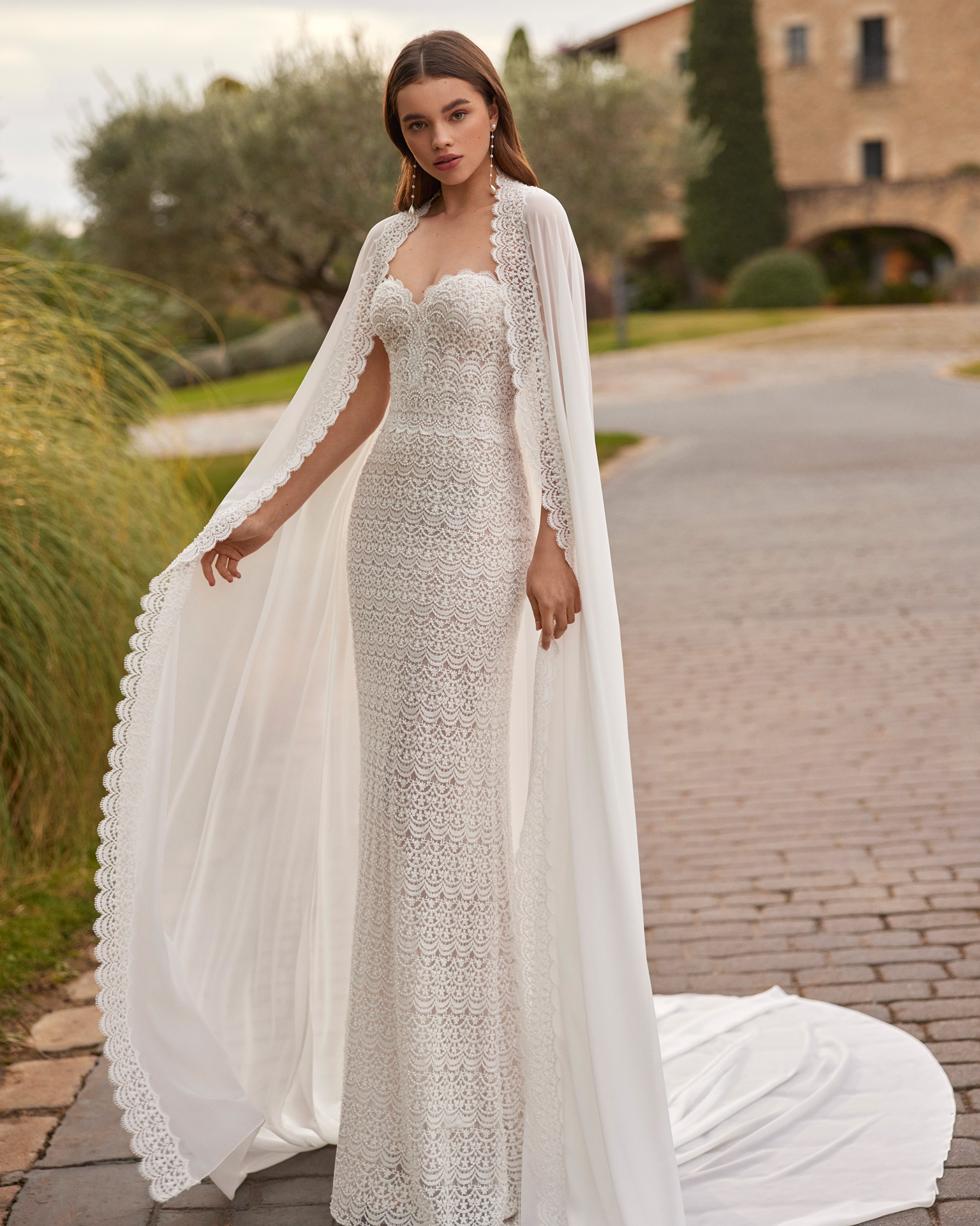 Vestido de noiva comprido, de corte sereia e estilo sexy, confecionado com renda e brilhantes. Apresenta um decote coração, costas corset e uma capa de georgette com aplicações de renda.