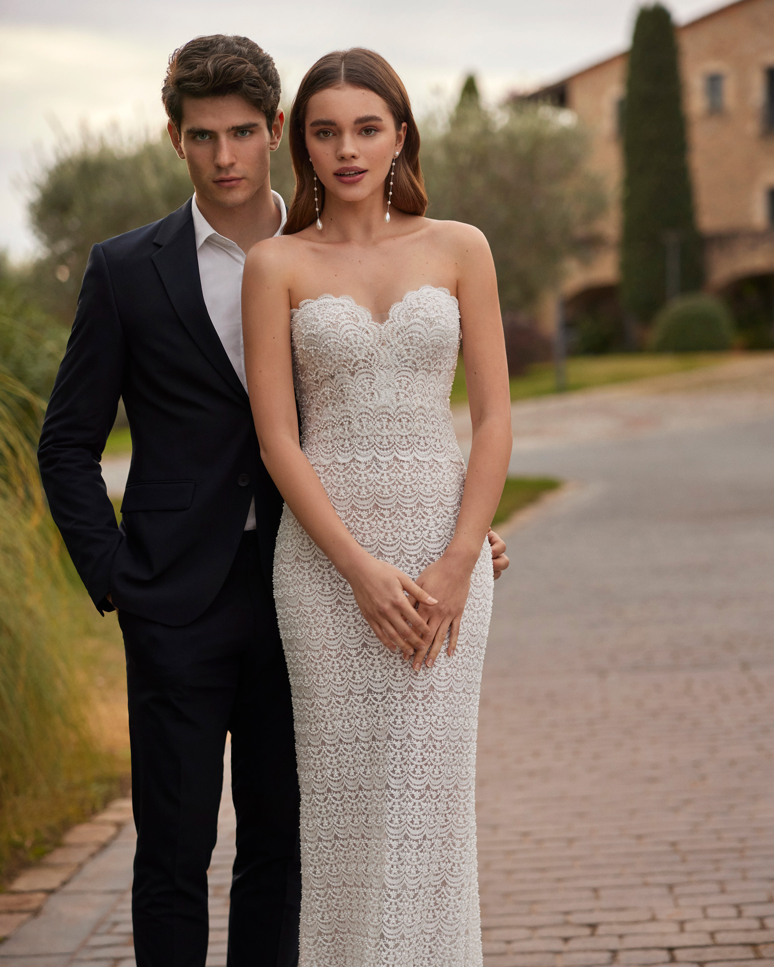 Vestido de noiva comprido, de corte sereia e estilo sexy, confecionado com renda e brilhantes. Apresenta um decote coração, costas corset e uma capa de georgette com aplicações de renda.
